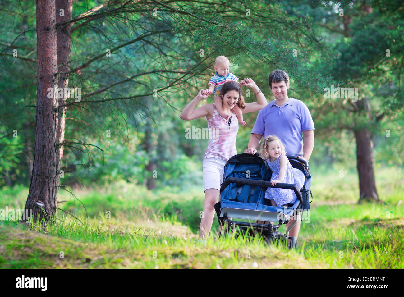 Active family, young parents and their two little children, hiking together in pine forest pushing an all terrain twin stroller Stock Photo