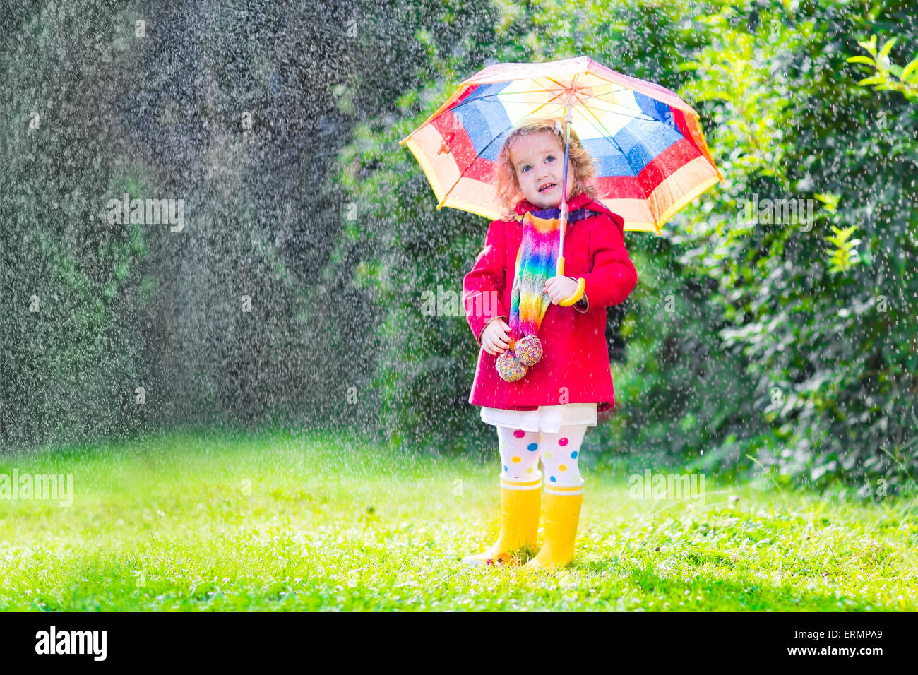 Funny cute toddler girl wearing red waterproof coat and yellow rubber boots holding colorful umbrella playing in garden by rain Stock Photo