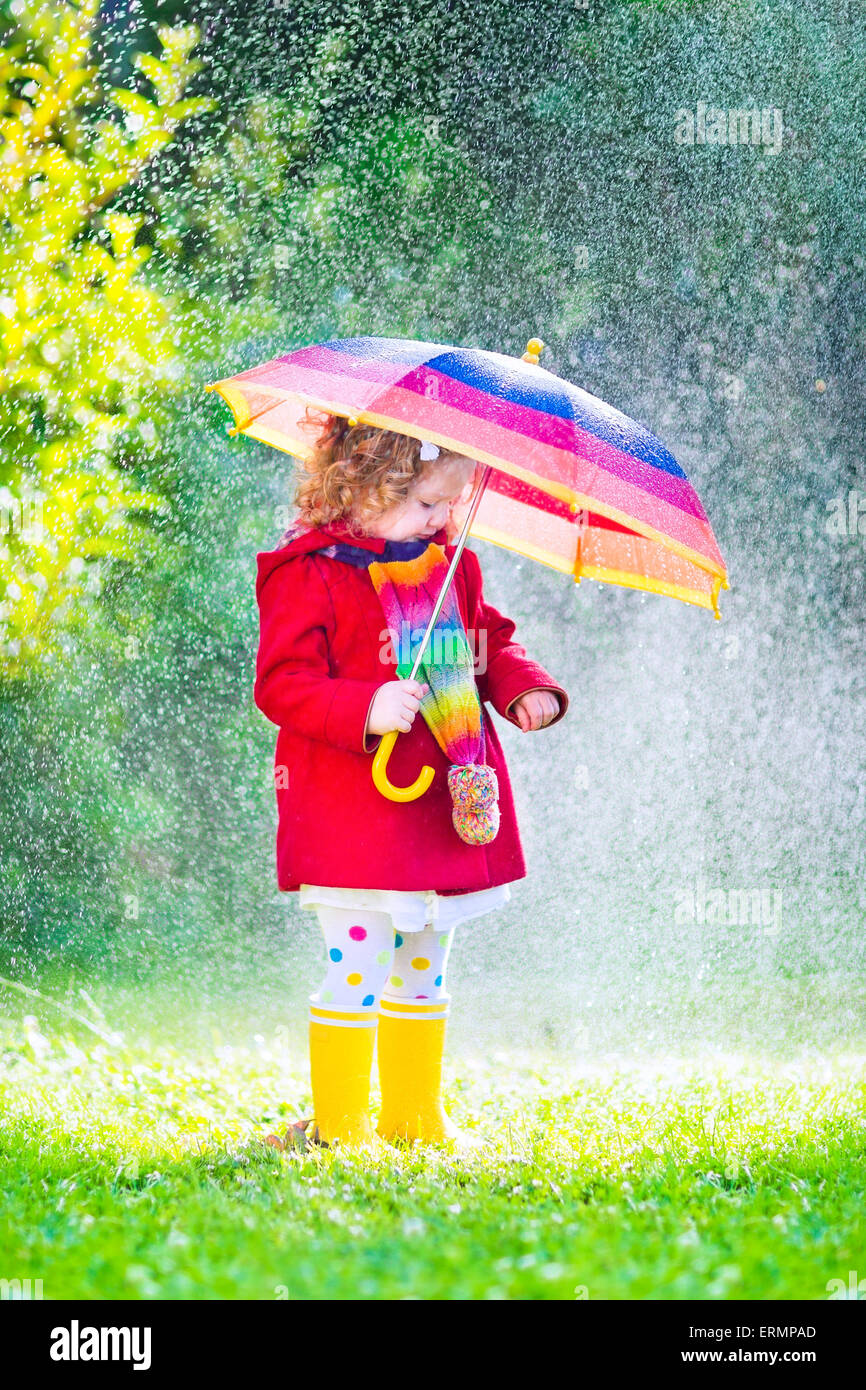 Funny cute toddler girl wearing red waterproof coat and yellow rubber boots holding colorful umbrella playing in garden by rain Stock Photo
