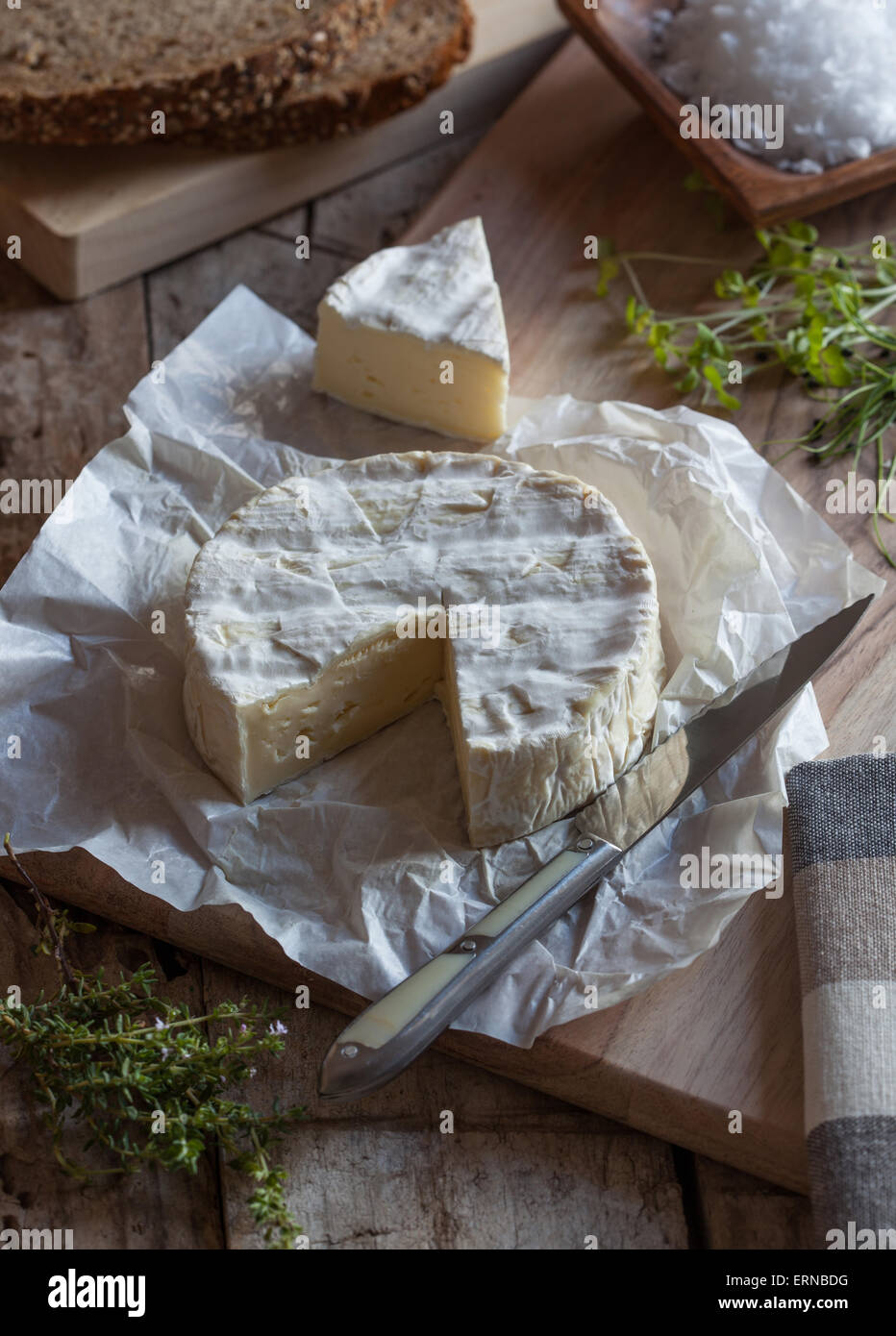 A piece of camembert cheese on a cutting board with a knife and herbs