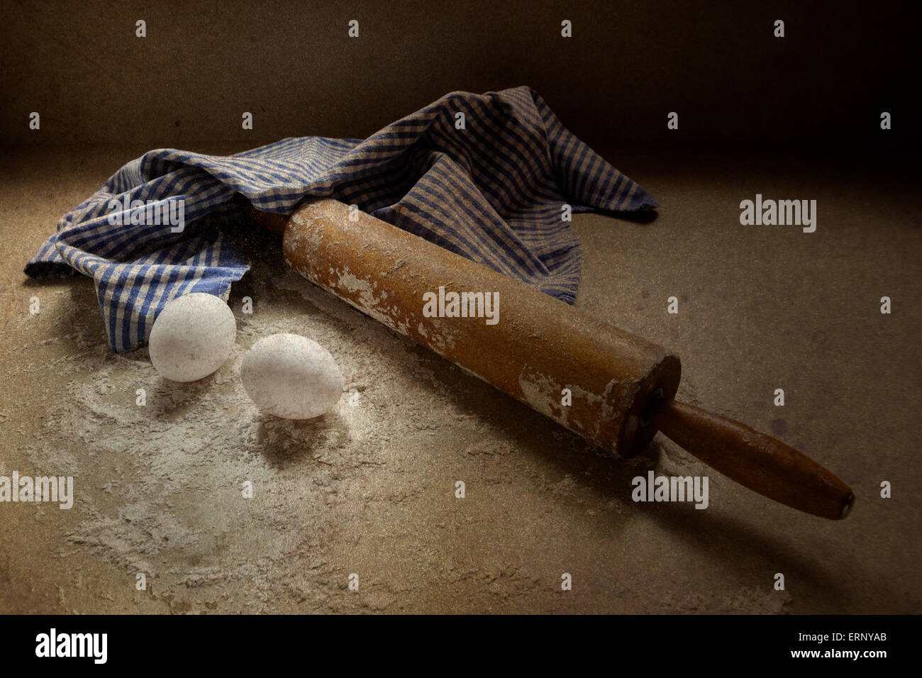 An antique rolling pin sits on a floured surface with two eggs and a checkered blue and white towel. Stock Photo