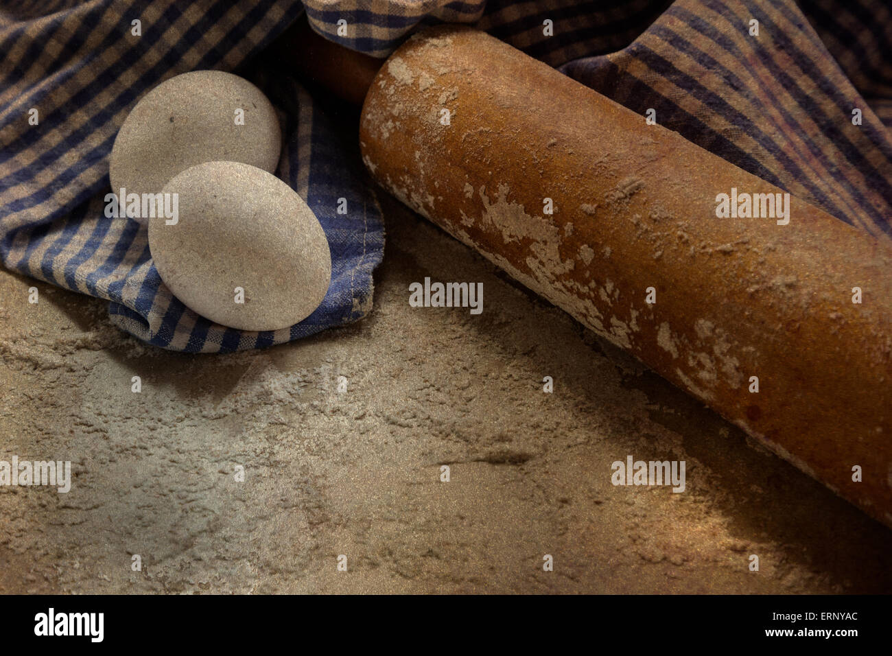 An antique rolling pin sits on a floured surface with two eggs and a checkered blue and white towel. Stock Photo