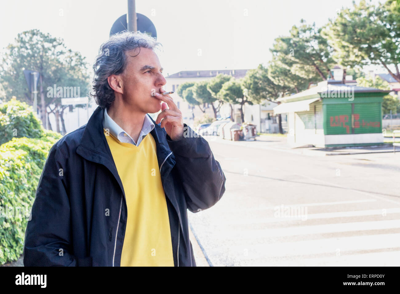 Handsome middle-aged man with salt pepper hair, medium hair, dressed in casual clothing with yellow sweater, slacks blue and yellow sneakers in green outdoors: he is smoking a cigarette Stock Photo