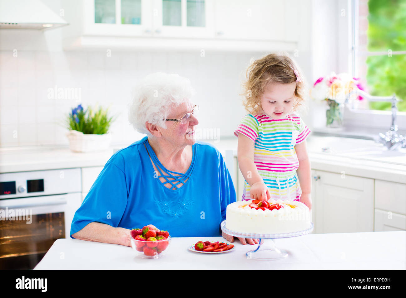 Happy senior lady, loving grandmother, baking a homemade strawberry cake with her granddaughter in a white modern kitchen Stock Photo