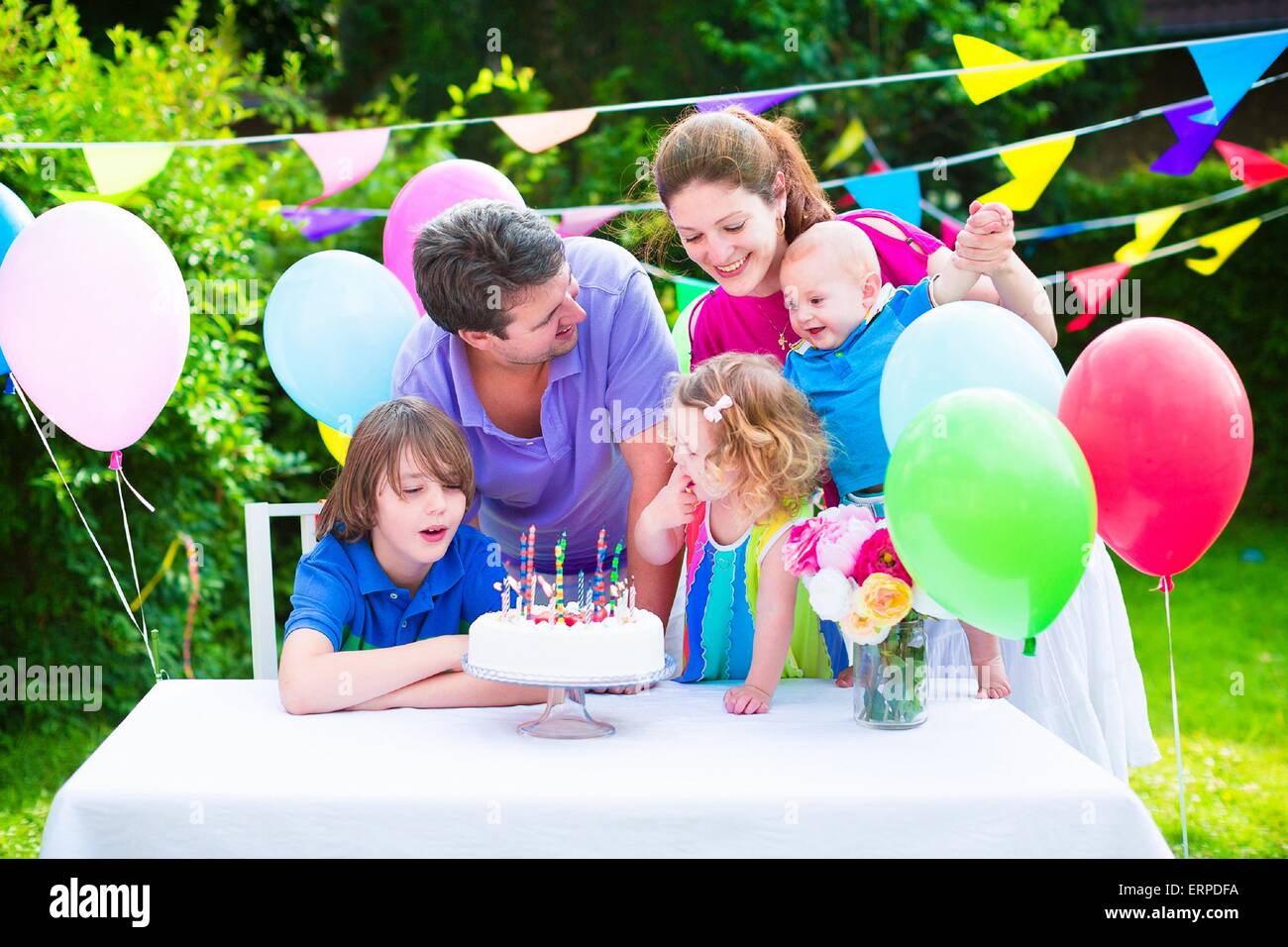 Happy big family with three kids enjoying birthday party with cake blowing candles in garden decorated with balloons and banners Stock Photo