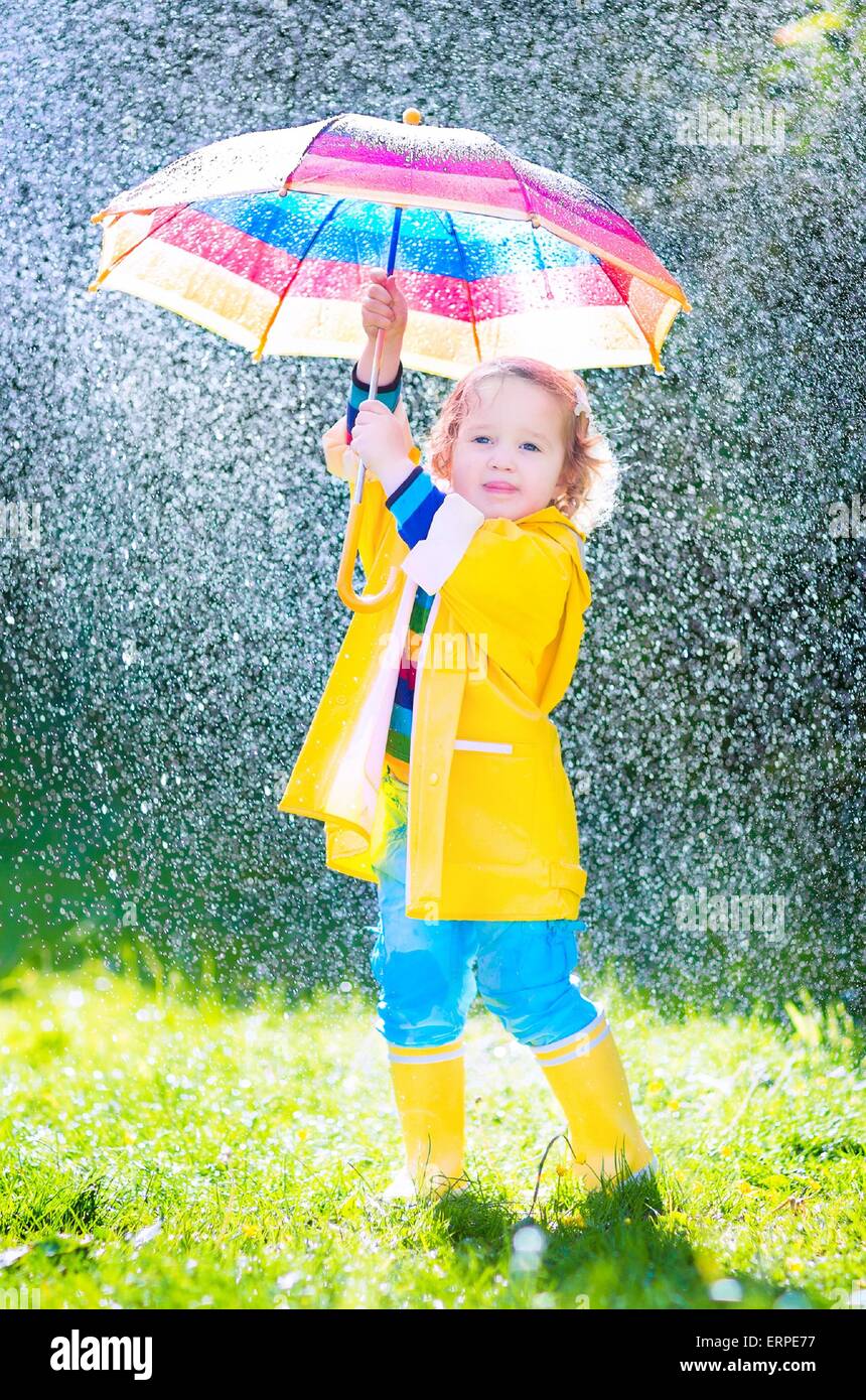 Funny cute curly toddler girl wearing yellow waterproof coat and boots holding colorful umbrella playing in the garden by rain Stock Photo