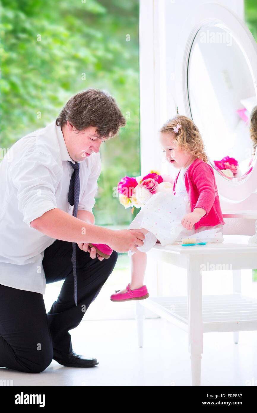 Young happy father helping his little daughter to put on a shoe, at a white dresser with mirror in a bedroom with a big window Stock Photo