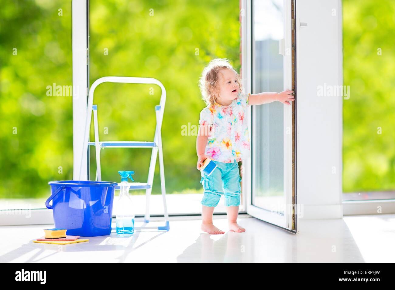 Cute laughing curly toddler girl washing a big window with a squeegee in beautiful white living room with door into the garden Stock Photo