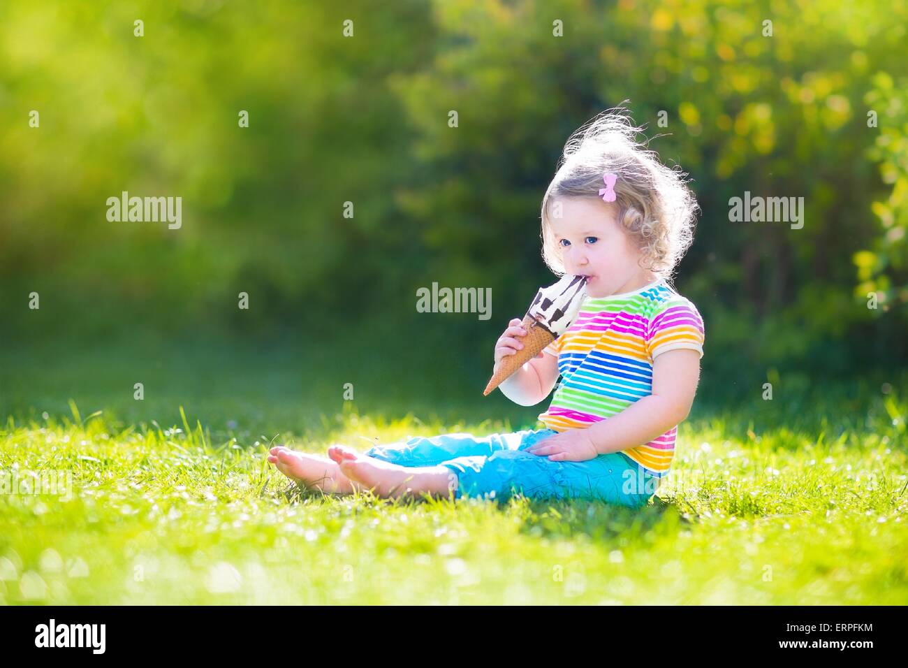 Funny toddler girl with curly hair wearing colorful summer shirt and blue jeans sitting on green lawn eating ice cream cone Stock Photo