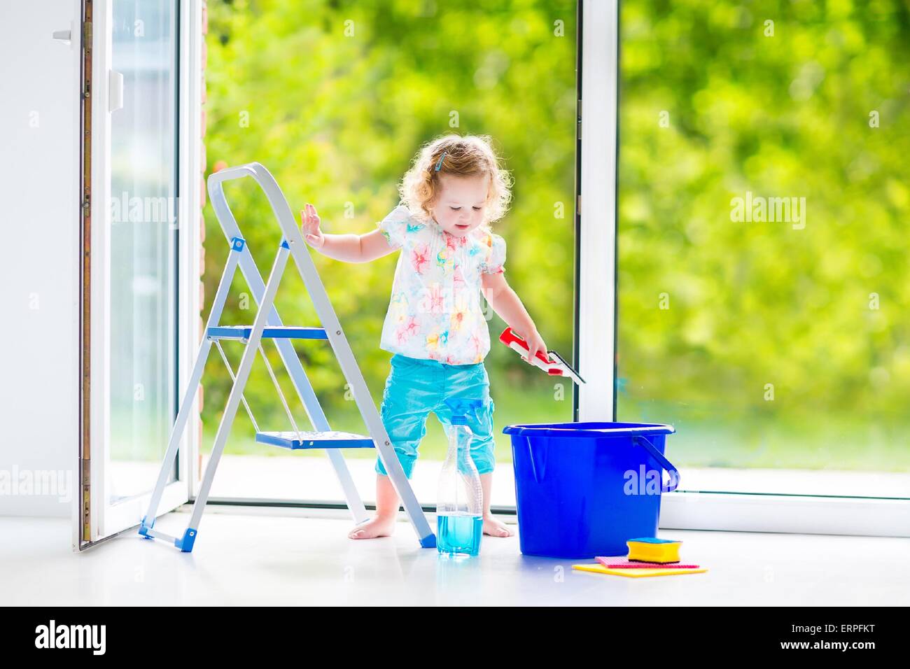 Cute laughing curly toddler girl washing a big window with a squeegee in beautiful white living room with door into the garden, Stock Photo