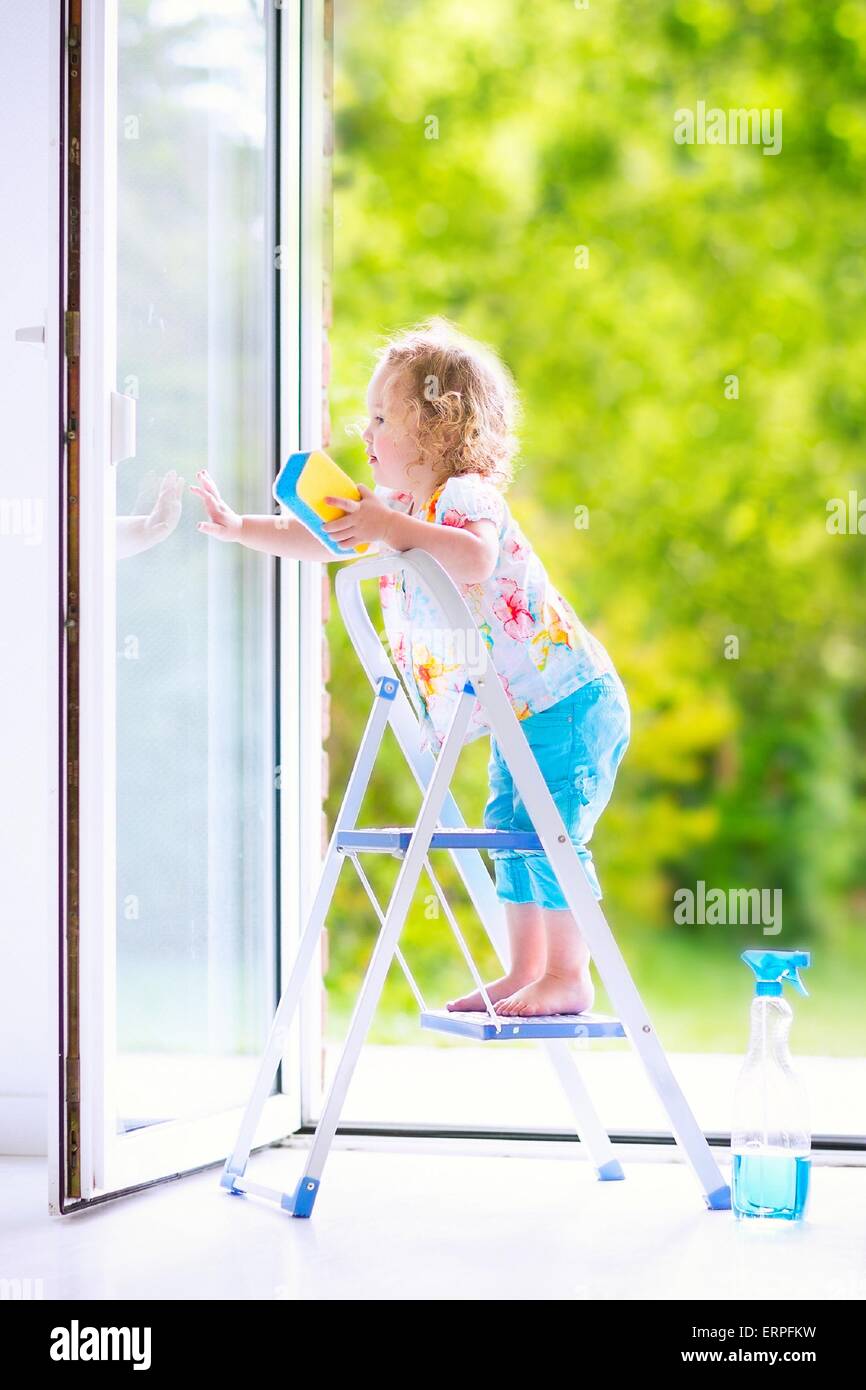 Cute laughing curly toddler girl washing a big window with a squeegee in beautiful white living room with door into the garden, Stock Photo