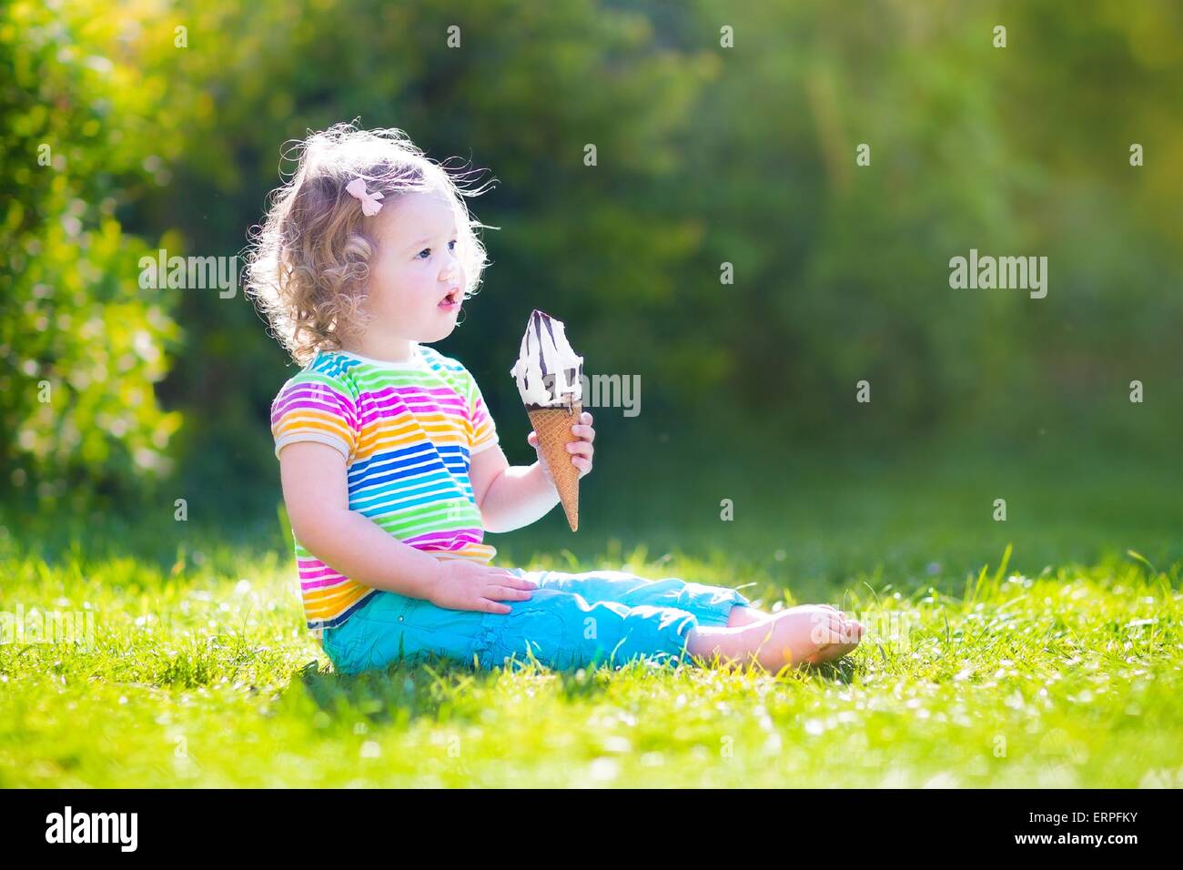 Funny toddler girl with curly hair wearing colorful summer shirt and blue jeans sitting on green lawn eating ice cream cone Stock Photo