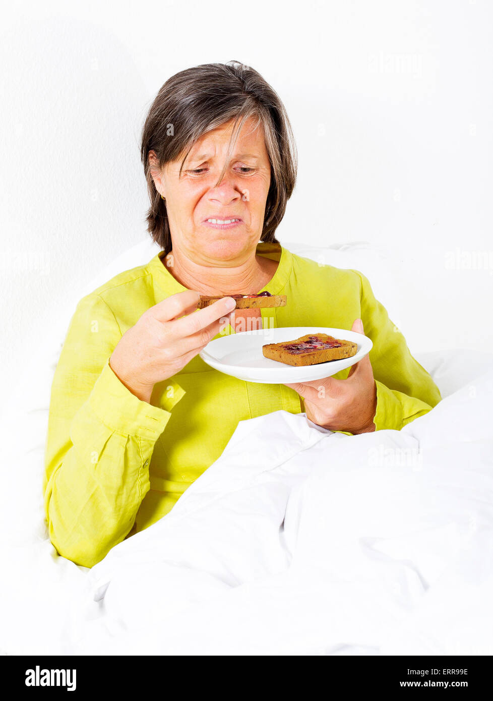 woman in her fifties sitting in her bed with a plate of toast and looking upset Stock Photo