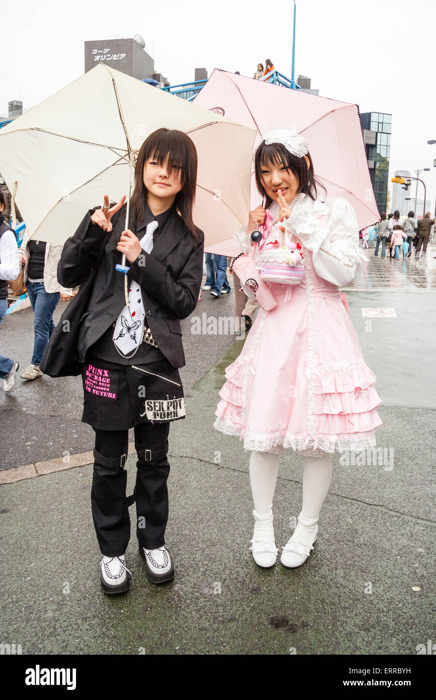 Two young Japanese women in Harajuku, posing in the street in the rain under umbrellas. One in pink, goth lolita maid costume, the other in black. Stock Photo
