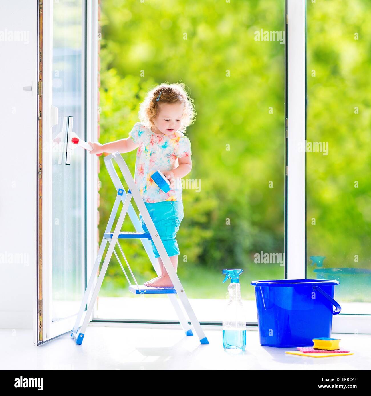 Cute laughing curly toddler girl washing a big window with a squeegee in beautiful white living room with door into the garden Stock Photo