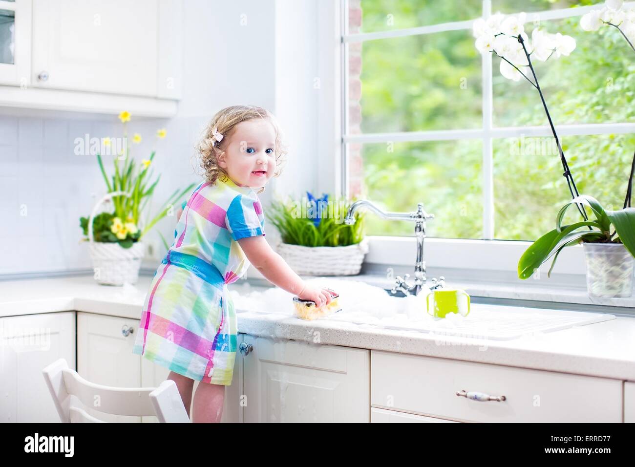 Cute curly toddler girl in a colorful dress washing dishes, cleaning with a sponge and playing with foam in the sink in kitchen Stock Photo