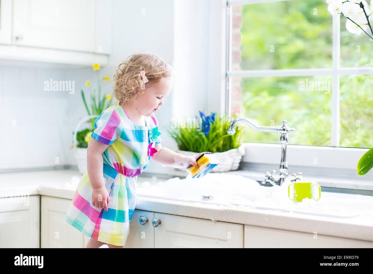 Cute curly toddler girl in colorful dress washing dishes, cleaning with a sponge and playing with foam in the sink in kitchen Stock Photo