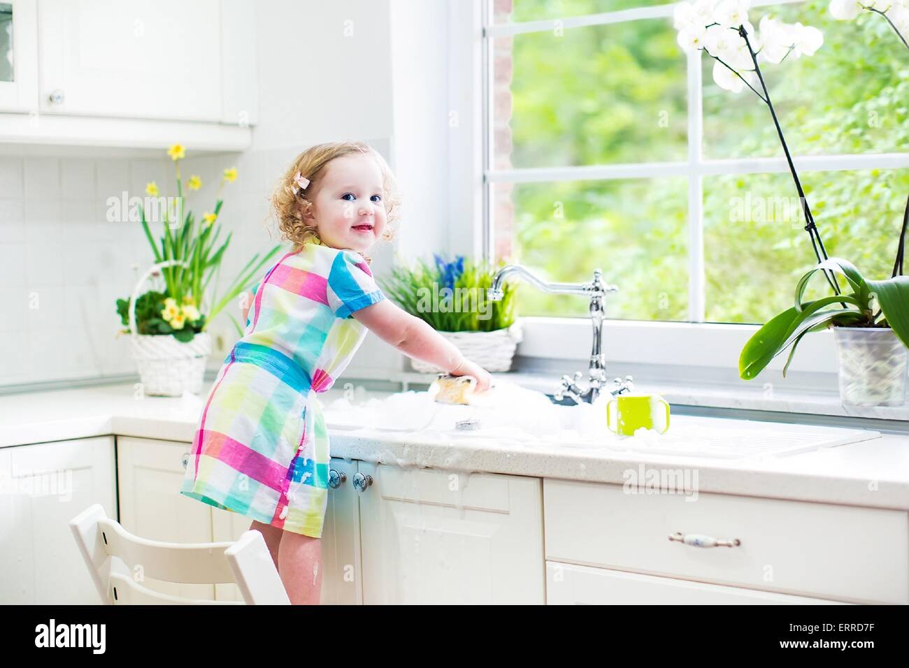 Cute curly toddler girl in a colorful dress washing dishes, cleaning with a sponge and playing with foam in the sink in kitchen Stock Photo