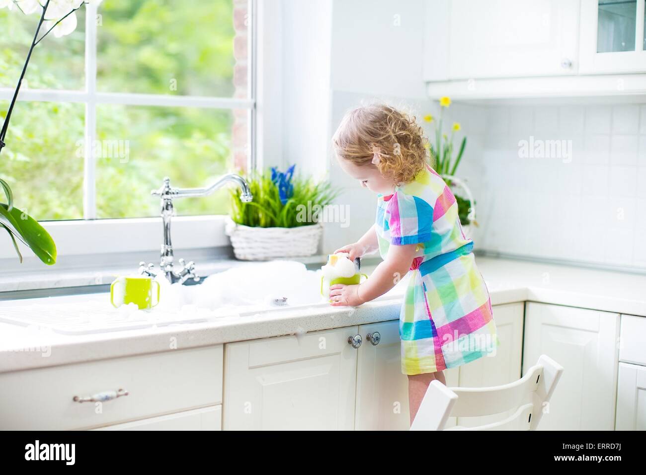 Child washing dishes. Kids wash plates and cups. Little girl helping in the kitchen playing with water and foam in a white sink Stock Photo