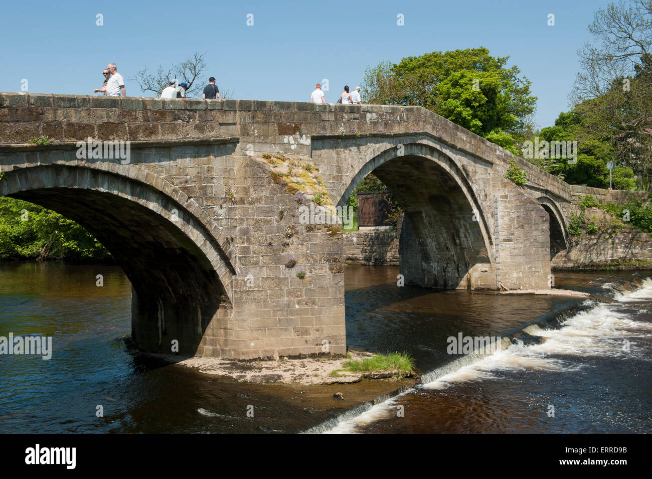 People crossing scenic River Wharfe on old stone packhorse bridge over flowing water & small weir - Old Bridge, Ilkley, West Yorkshire, England, UK. Stock Photo