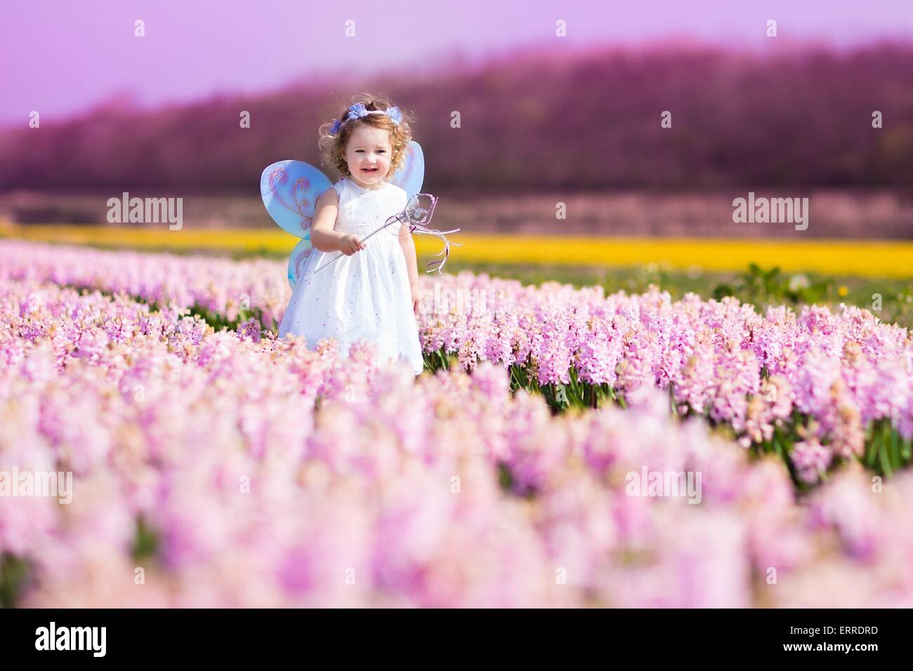 Portrait of an adorable toddler girl in a magic fairy costume and flower crown in her curly hair playing with a wand in field Stock Photo