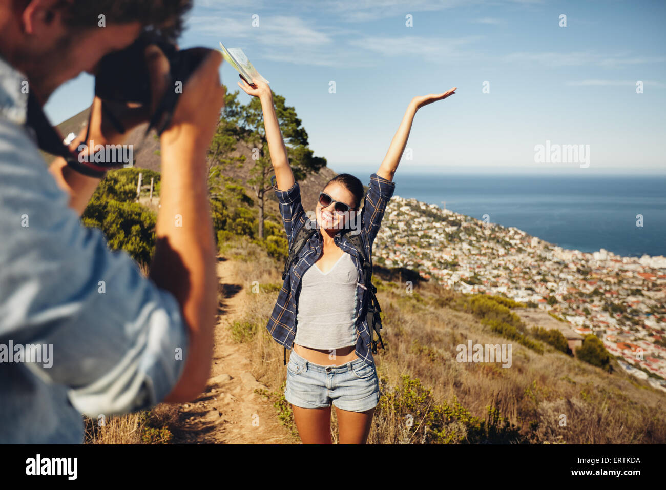 Happy young woman posing with her arms raised to her boyfriend taking her photos with digital camera. Summer vacation fun, young Stock Photo