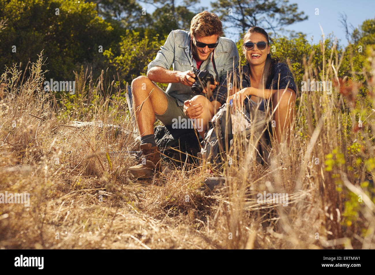 Outdoor shot of young couple on hiking trip taking a break. Woman smiling while man looking at his camera. Stock Photo
