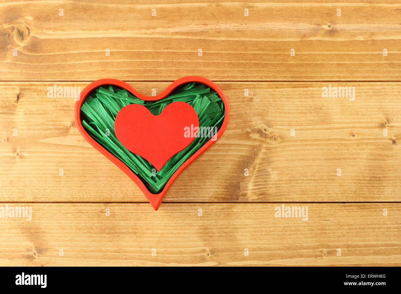 Red painted wooden heart enclosed with green paper raffia strips in red box on wooden background Stock Photo