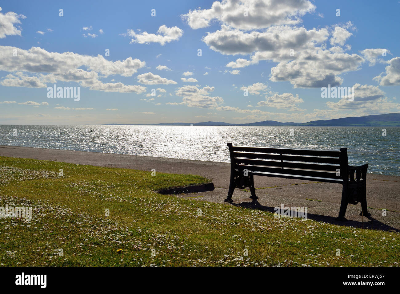 looking out into the solway coast on a bright sunny afternoon with a seat in the foreground blue sky and white fluffy clouds Stock Photo