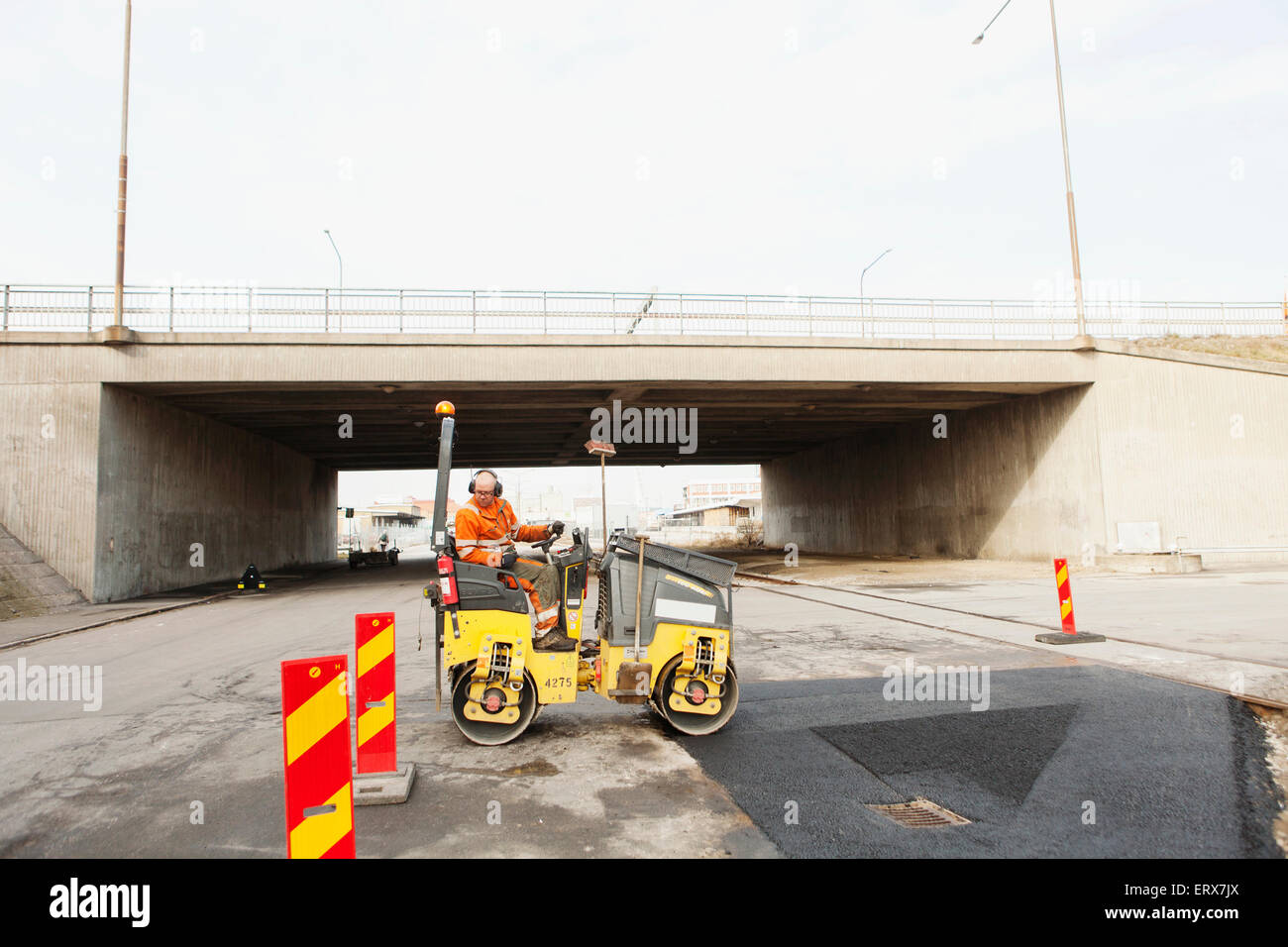 Manual worker driving steamroller on tar by bridge - Stock Image