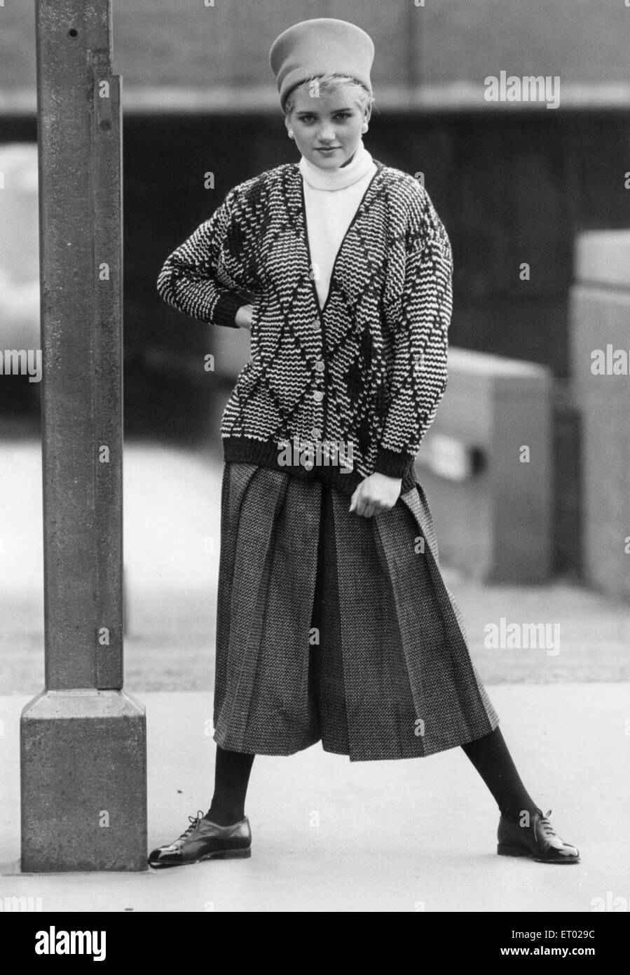 1980s Women's Fashion: Our model wears. Diamond pattern cardigan over a white turtle neck jumpers, full length pleated skirt, black leggings and black flat shoes and hat  6th October 1986 Stock Photo