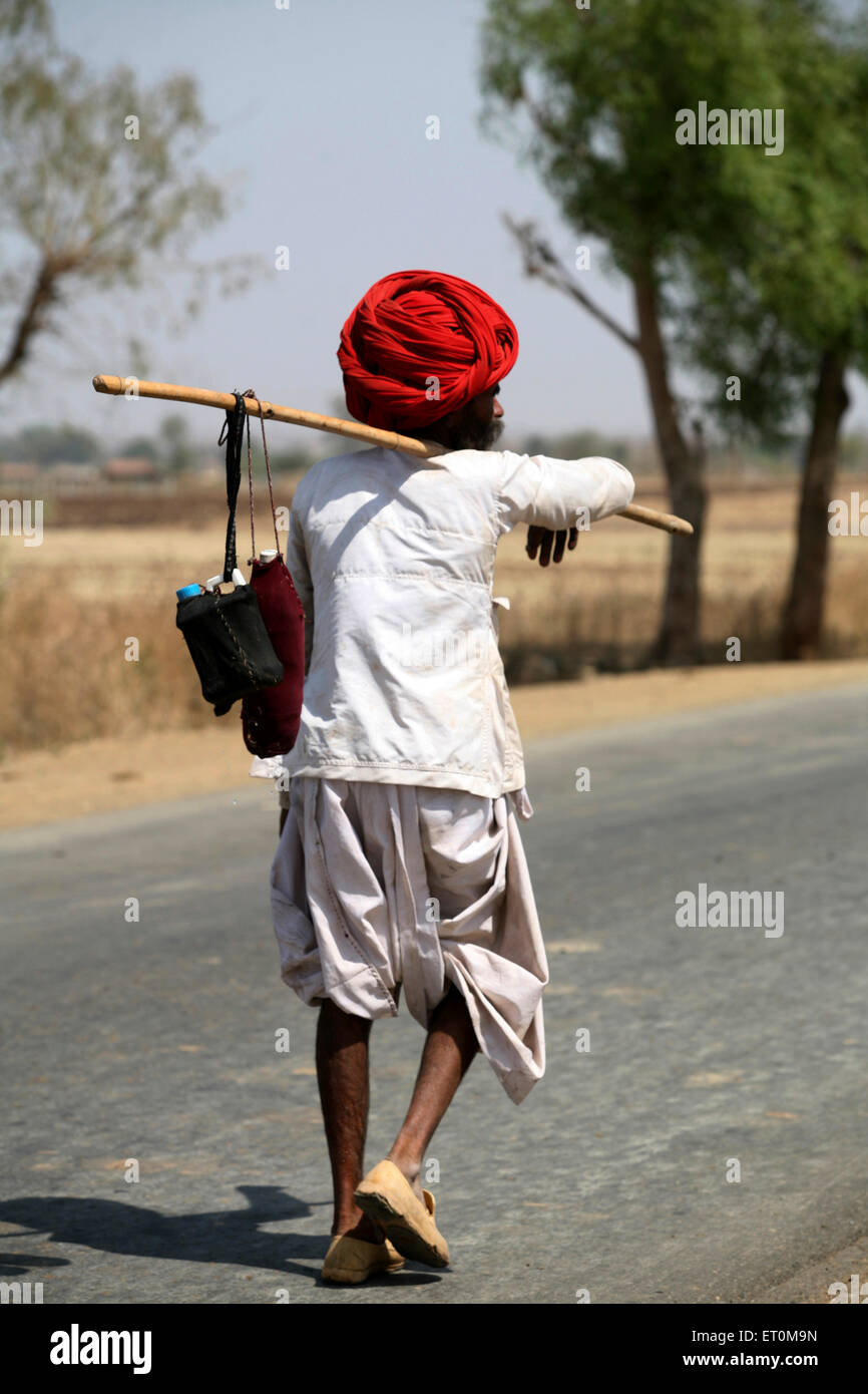 Nomad carrying set of bottles tied to his stick on his shoulders on Bhopal highway in Madhya Pradesh ; India Stock Photo