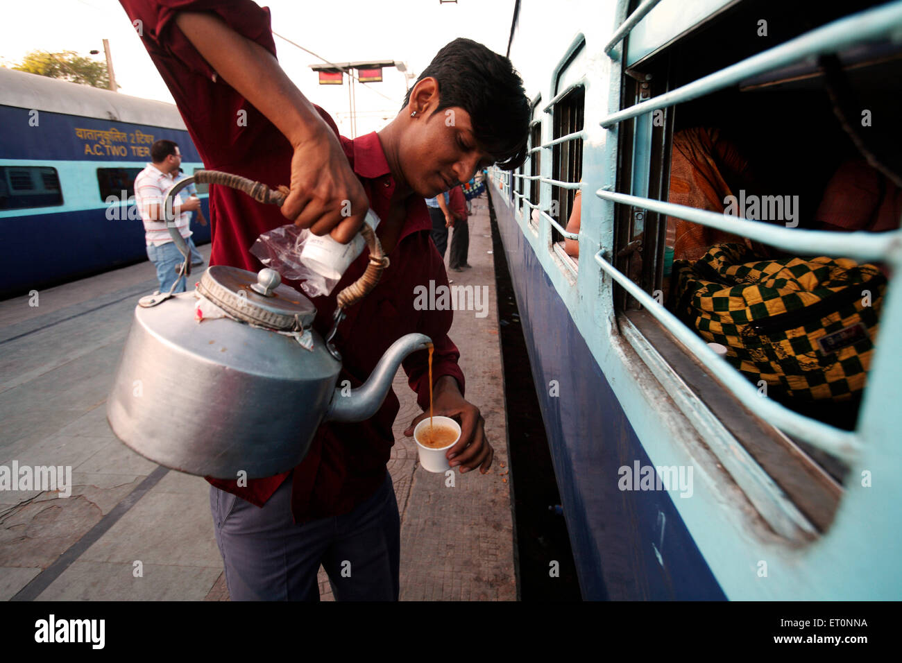 Indian railway tea vendor tea seller pouring tea from aluminium kettle into plastic cup for passengers in train at railway station in India Asia Stock Photo