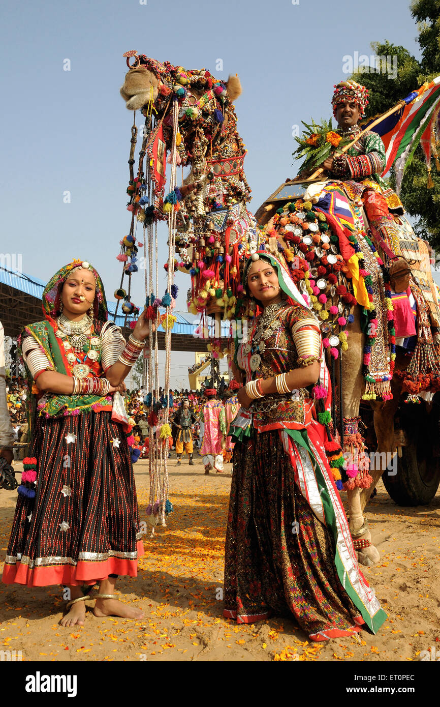 Girls in traditional jewellery and rajasthani costume standing in front of  decorated camel in Pushkar fair ; Rajasthan Stock Photo