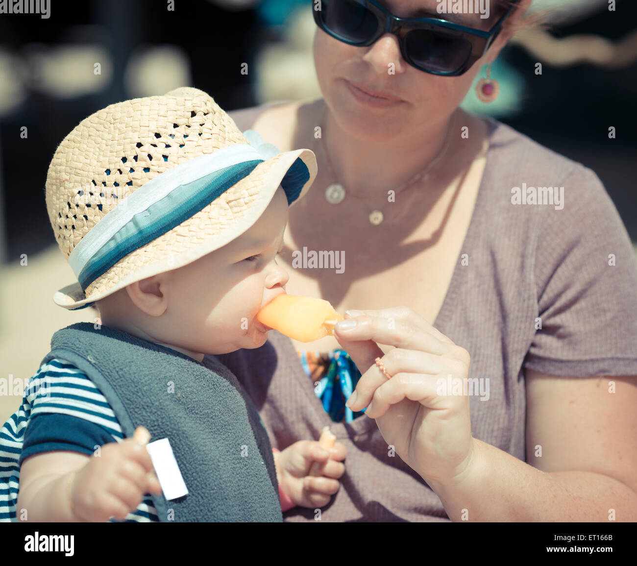 little baby girl sitting with her mother on the beach eating aan ice-cream with her hat on Stock Photo
