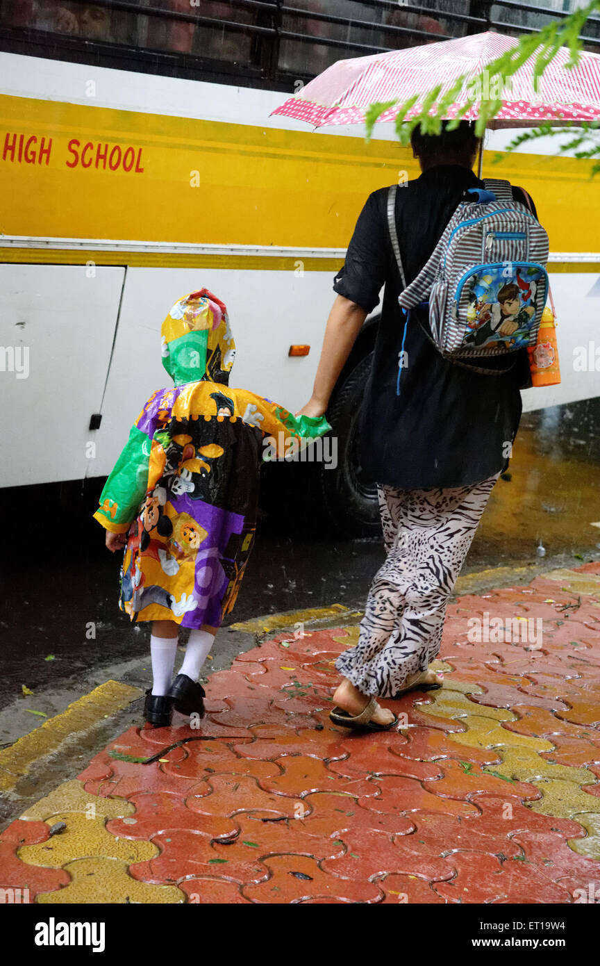 Mother with umbrella walking boy in raincoat to school bus in monsoon season rain India - MR#364 Stock Photo