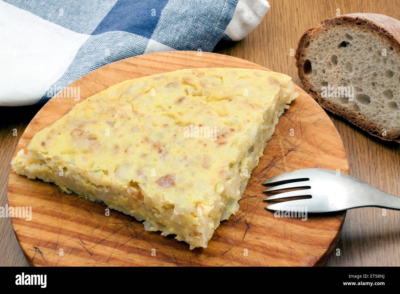 a piece of a spanish omelet over a wood board with a fork and a piece of cloth Stock Photo