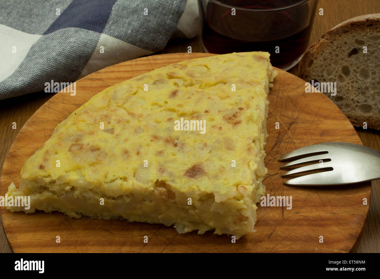 a piece of a spanish omelet over a wood board with a fork, a glass of wine and a piece of cloth Stock Photo