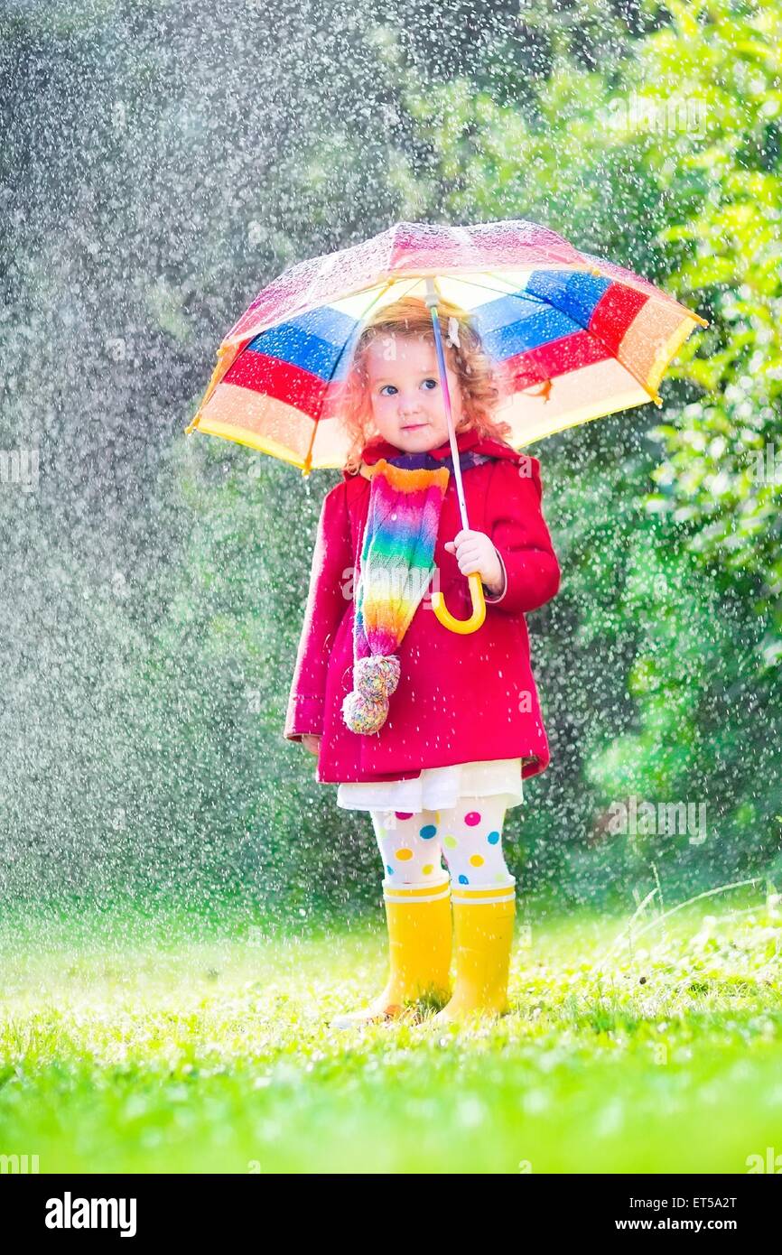 Funny cute curly toddler girl wearing red waterproof coat and yellow rubber boots holding colorful umbrella playing in garden Stock Photo