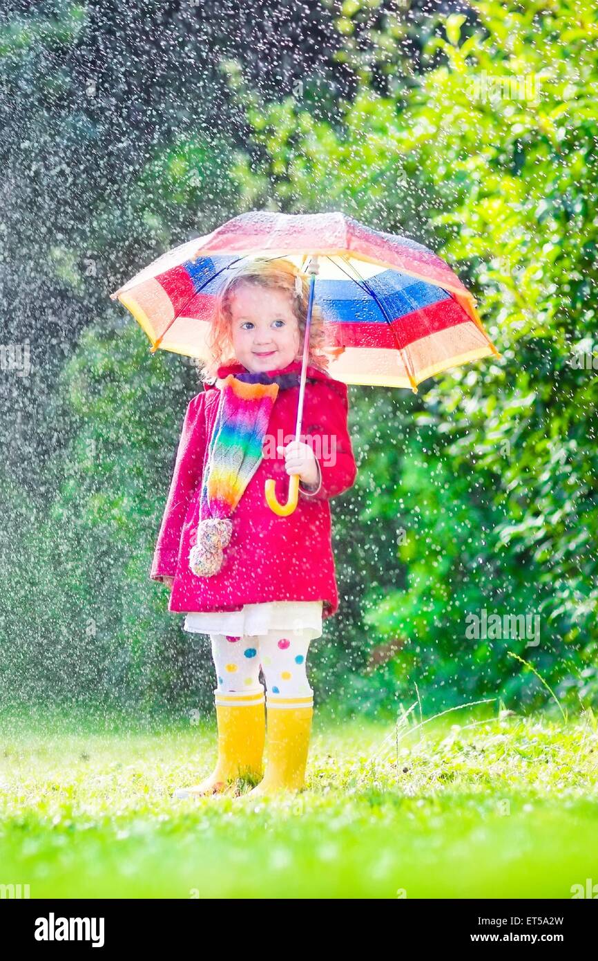 Funny cute curly toddler girl wearing red waterproof coat and yellow rubber boots holding colorful umbrella playing in garden Stock Photo