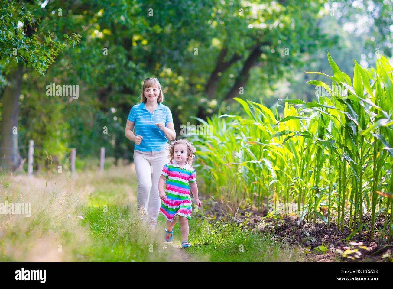 Happy family enjoying a summer day in the forest, active young woman and a cute toddler girl running next to a corn field Stock Photo