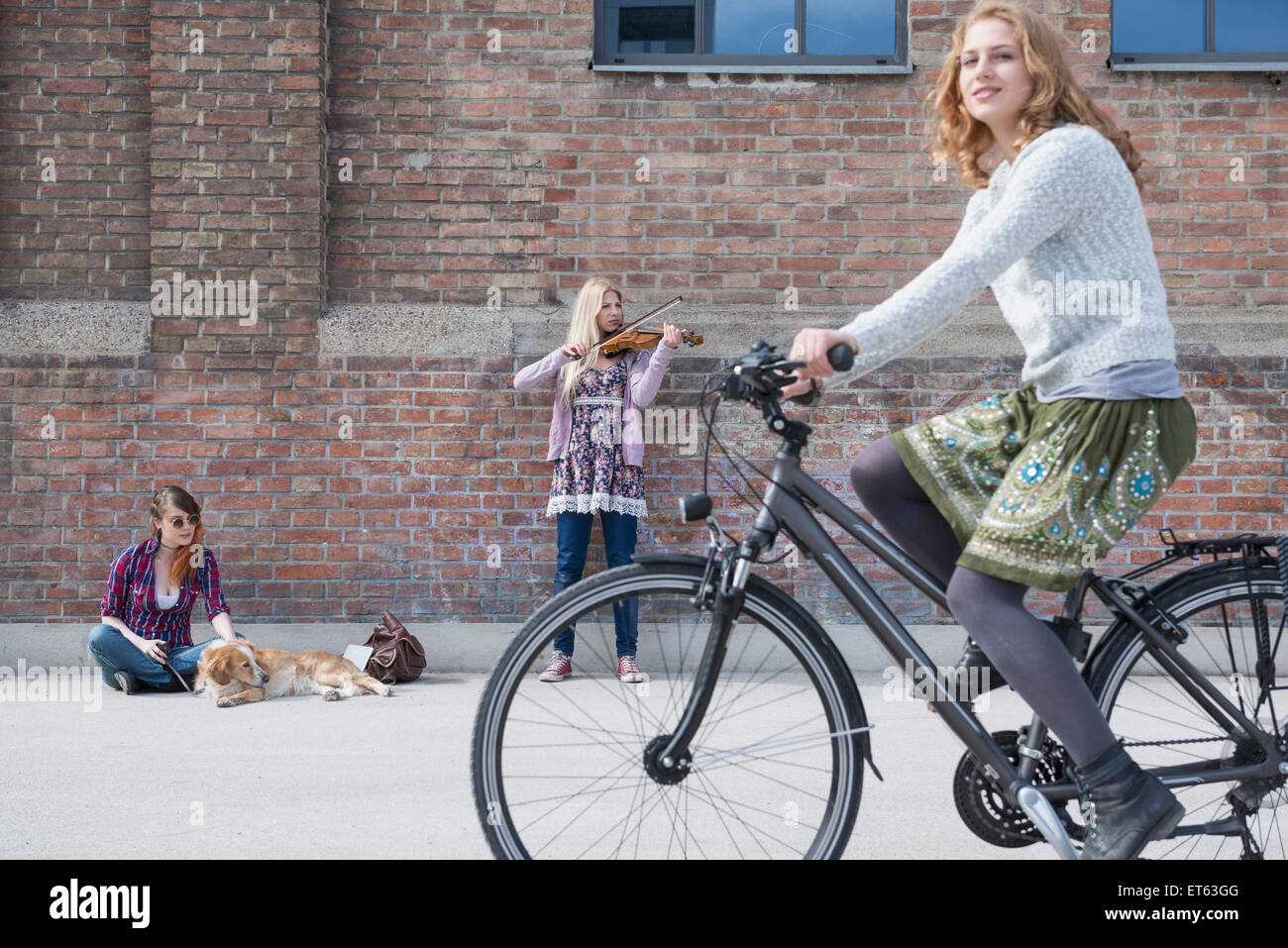 Woman riding bicycle on street with teenage girl playing violin in background, Munich, Bavaria, Germany Stock Photo