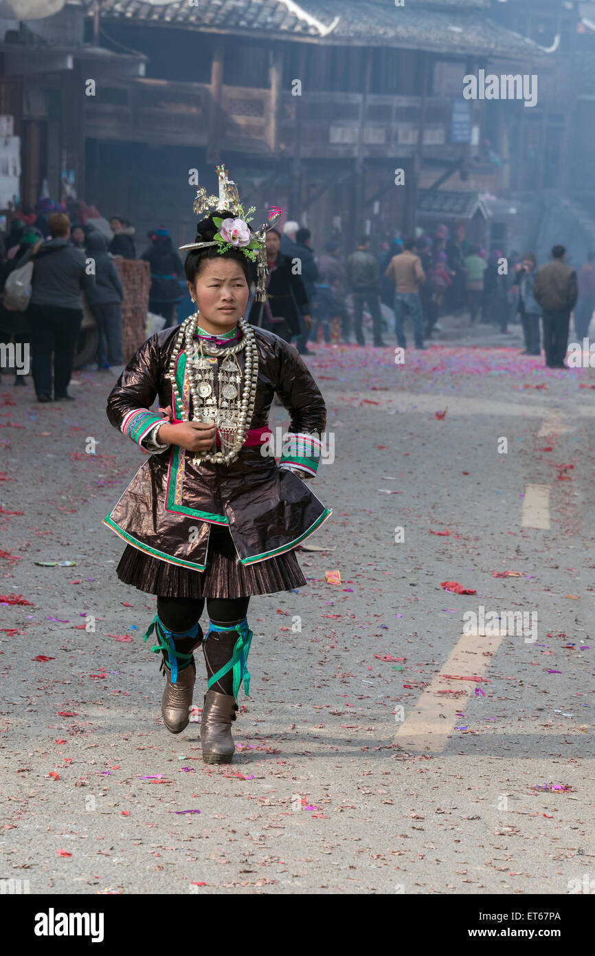 Woman in ethnic Dong attire hurrying down a street littered with firecracker remnants, Huanggang Dong Village, Guizhou Province, Stock Photo