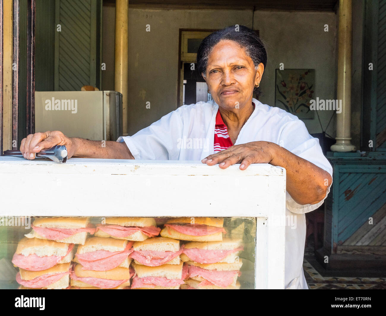 A older Afro-Cuban female sandwich vendor leans on her display case holding sandwiches and other foods while holding tongs. Stock Photo