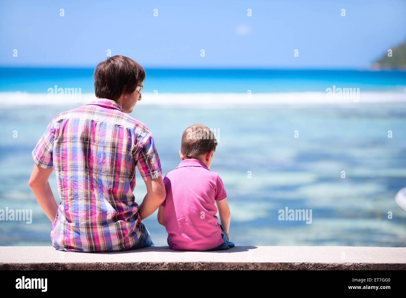 Vater sitzt mit seinem Sohn auf einer Mauer und blickt auf das Meer, Rueckansicht | father with his son sitting on wall and view Stock Photo