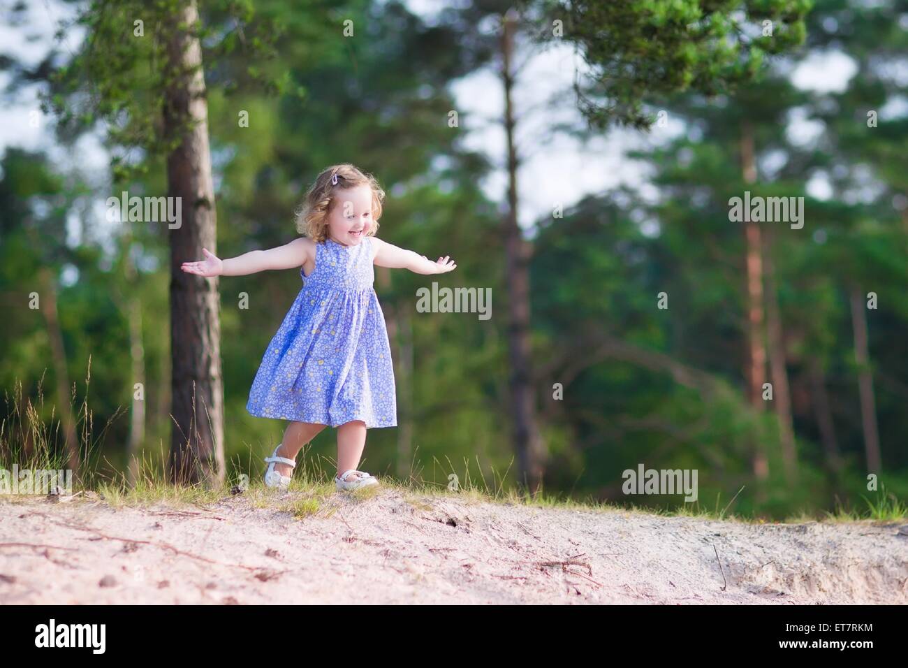Adorable child, little curly toddler girl in a blue summer dress running and playing on sand dunes in beautiful pine wood forest Stock Photo
