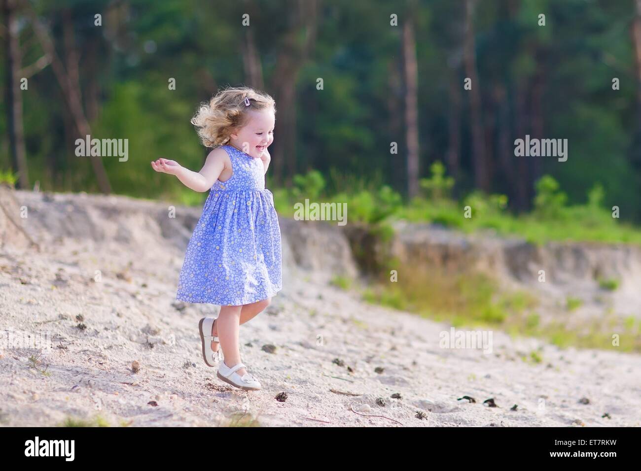 Adorable child, little curly toddler girl in a blue summer dress running and playing on sand dunes in a beautiful pine wood fore Stock Photo