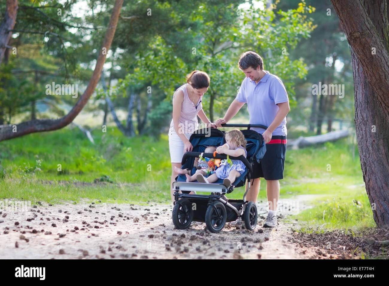 Active family, young parents and their two little children hiking together in a pine forest pushing an all terrain twin stroller Stock Photo