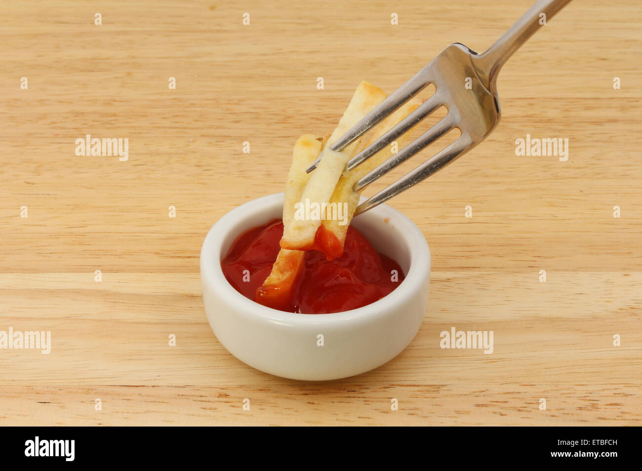 French fries on a fork dipping in tomato ketchup on a wooden board