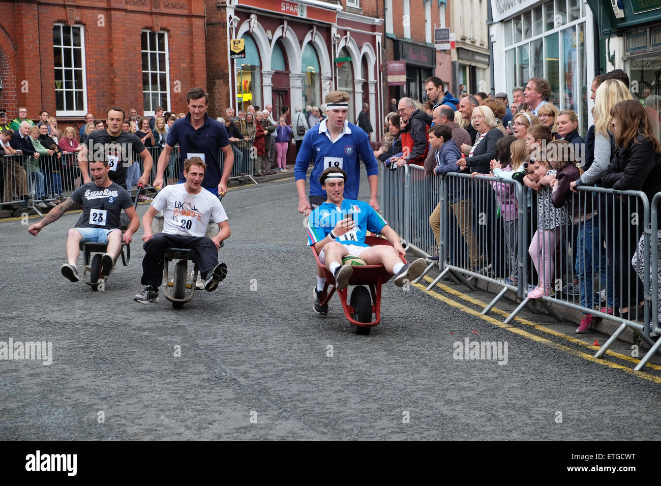 Kington, Herefordshire, UK. 13th June, 2015. Kington Festival Wheelbarrow Race competitors in this years 39th Wheelbarrow Race raced around town stopping to drink at each pub en route. Here three teams race through the town centre past the crowds. Stock Photo